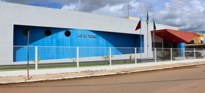 Imagem Fachada do prédio da Justiça Federal em Sousa. A estrutura é moderna, com paredes brancas e uma grande faixa azul na parte frontal, decorada com três janelas redondas. À frente do prédio, há três mastros com bandeiras, e uma cerca branca delimita o terreno. 

