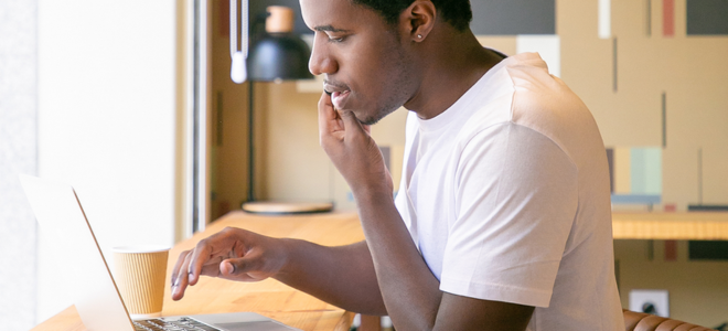 Imagem A foto mostra um homem jovem, com cabelo curto e pele escura, vestindo uma camiseta branca. Ele está sentado em um ambiente interno, concentrado enquanto usa um laptop em uma mesa de madeira. Uma xícara descartável de café está ao lado dele. Ao fundo, há uma parede com decoração geométrica e uma luminária preta moderna. A luz natural entra pela janela, iluminando a cena.