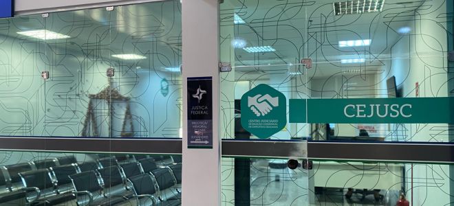 Imagem A imagem mostra a entrada do CEJUSC (Centro Judiciário de Solução Consensual de Conflitos) da Justiça Federal, com uma porta de vidro transparente decorada com linhas curvas em padrão repetido. No centro da porta há um adesivo verde em formato hexagonal com o ícone de duas mãos se cumprimentando, representando conciliação. Ao lado, em uma faixa verde horizontal, está escrito “CEJUSC” em letras brancas. À esquerda da porta, vê-se uma sala de espera com várias cadeiras metálicas enfileiradas. O ambiente é bem iluminado com luzes de teto e transmite uma atmosfera organizada e acolhedora.