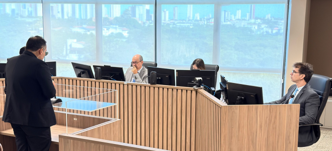 Imagem Três magistrados e a diretora de Secretaria da TR estão sentando atrás de uma mesa grande de madeira. De frente para eles e de costas para o público, um advogado, de pé, usa a tribuna para fazer uma sustentação oral. 
